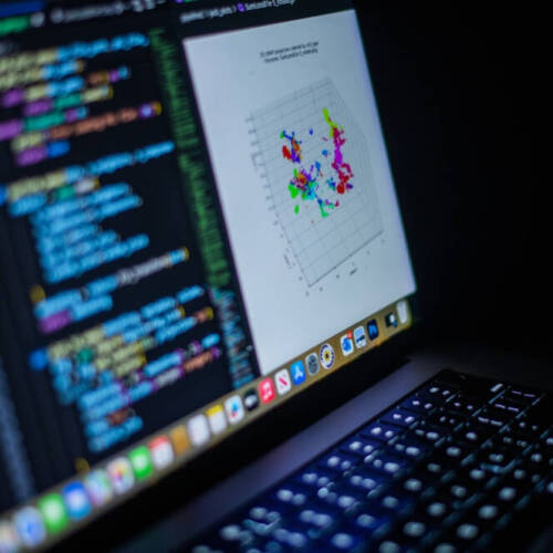 A laptop screen displaying colorful programming code on the left and a 3D scatter plot with multicolored data points on the right, set against a dark background.